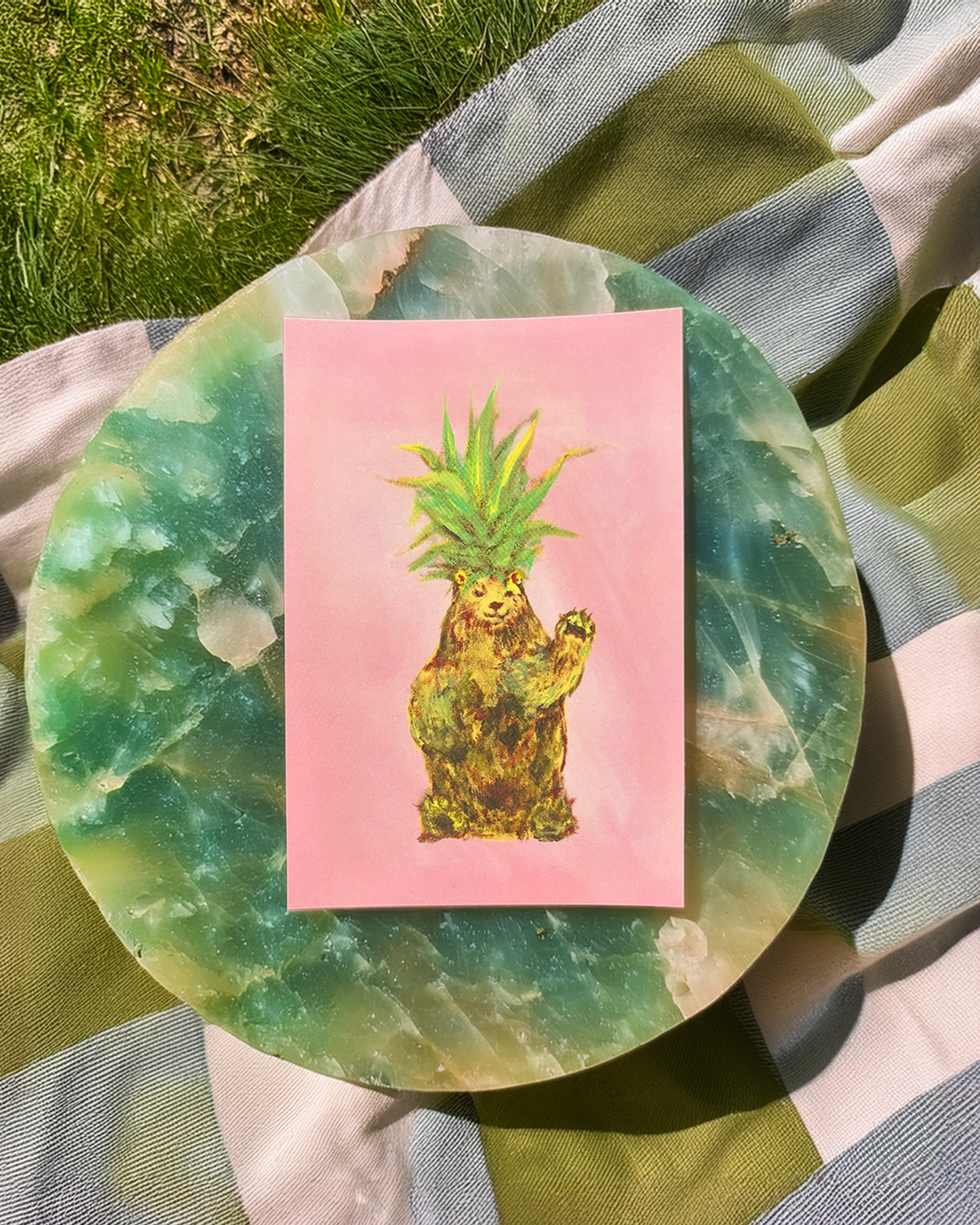 Card with a bear wearing a pineapple hat on a pink background, placed on a green marble surface outdoors.