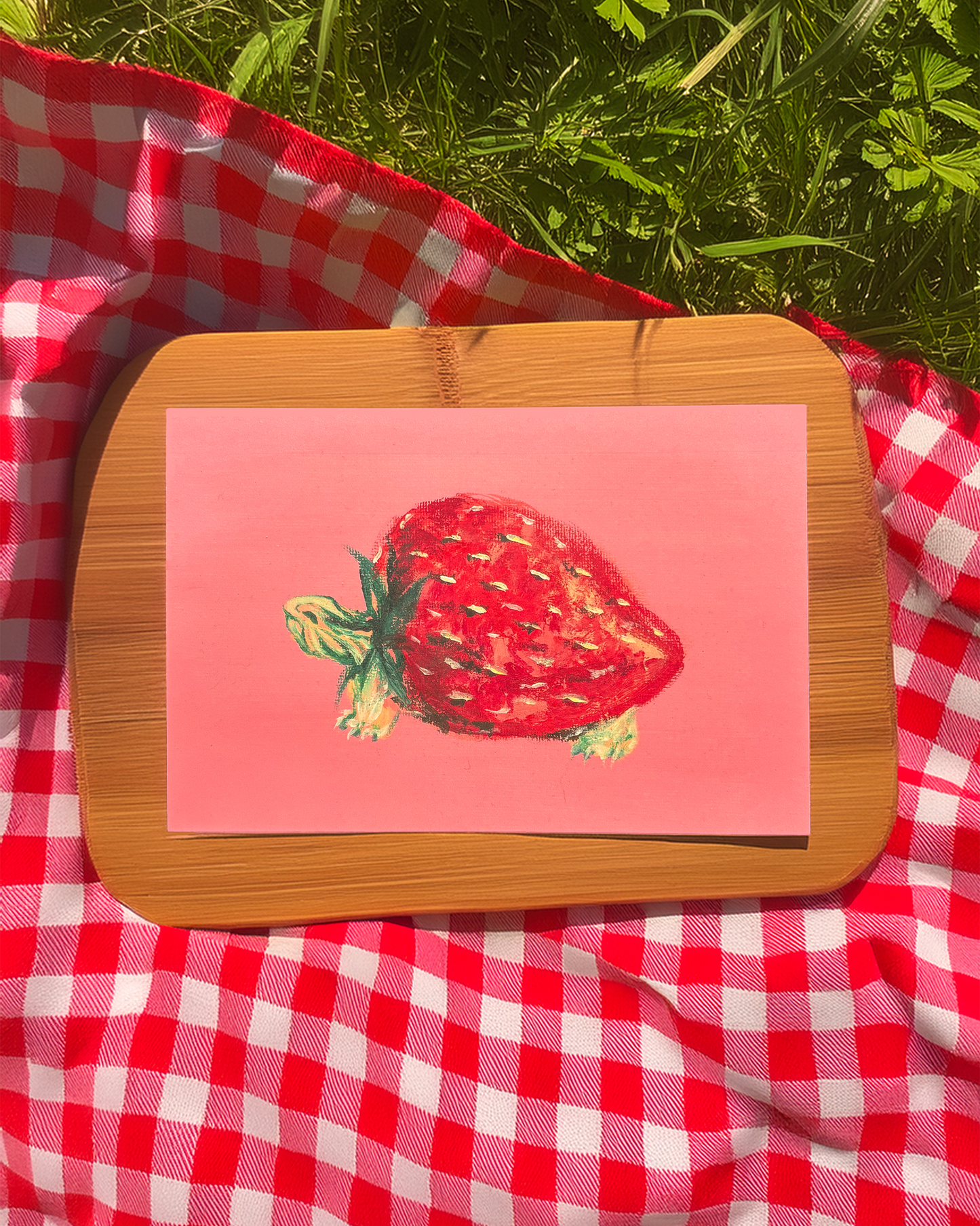 Strawberry illustration on a pink card with a wooden frame, placed on a red and white checkered cloth outdoors.